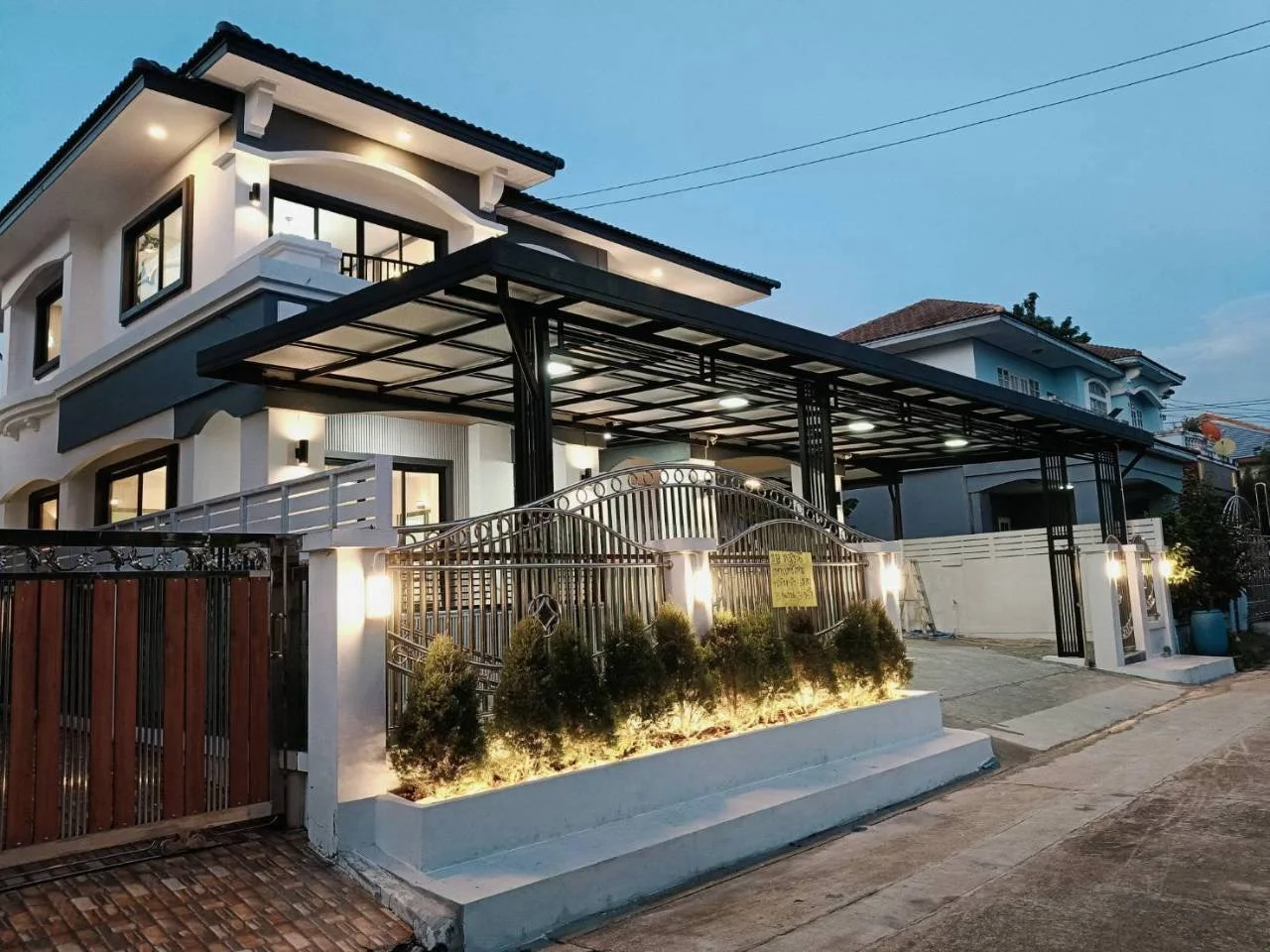 Front of the two-storey detached house at dusk, with a steel-frame carport and a planted bed along the fence